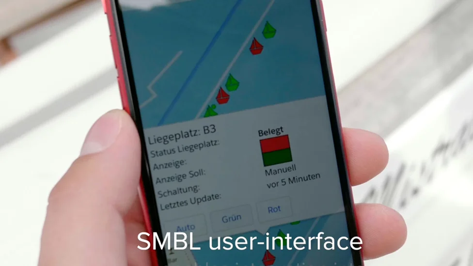 Ein in der Hand gehaltenes Handy. Auf dem Bildschirm ist eine Kartenansicht einer Marina zu sehen, mit eingezeichneten Liegeplätzen und Booten. Manche sind rot, manche grün markiert.